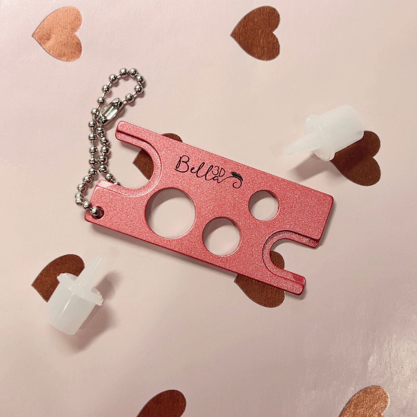 Pink metal Adhesive Nozzle Remover tool with keychain and two white plastic adhesive nozzles on a heart-patterned background