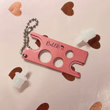Pink metal Adhesive Nozzle Remover tool with keychain and two white plastic adhesive nozzles on a heart-patterned background
