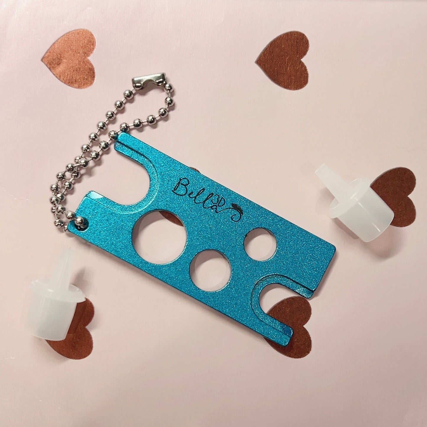 Blue Adhesive Nozzle Remover tool with keychain and white plastic nozzles on pink surface with heart shapes