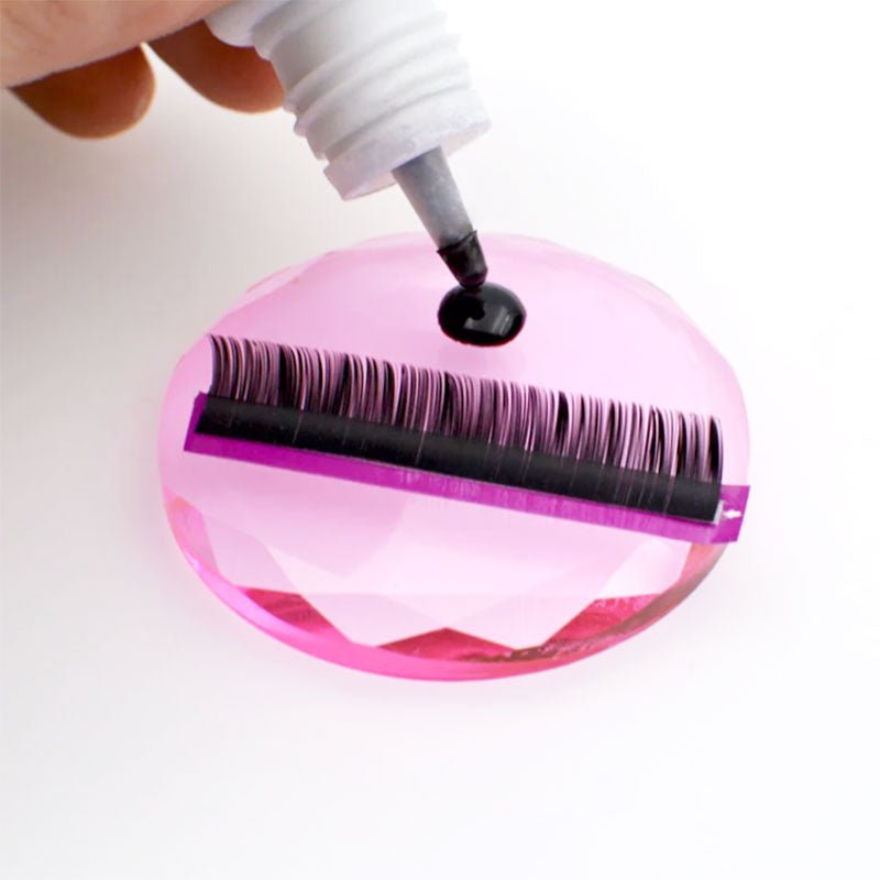 Close-up of a hand applying adhesive on Crystal Lash Glue Stones with eyelash extension supplies on top.