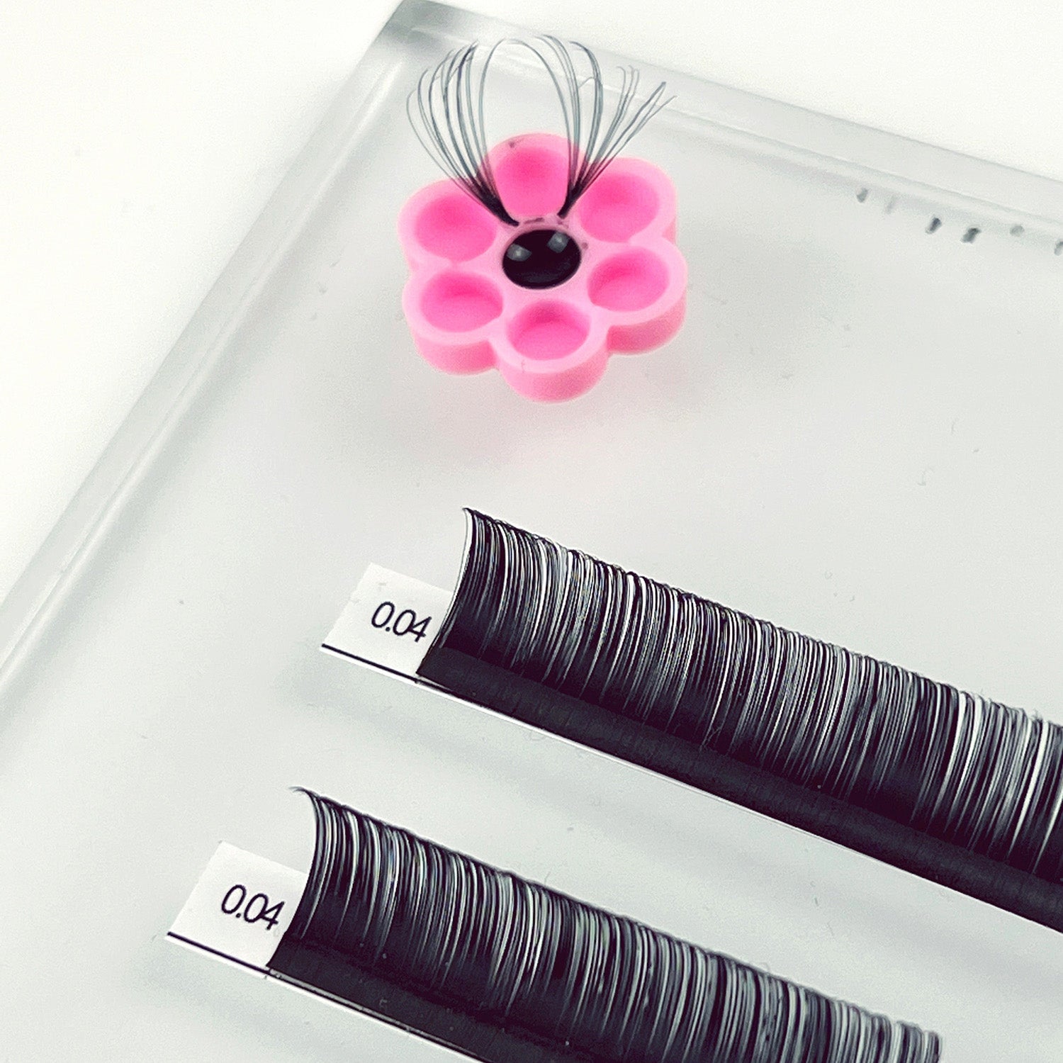 Pink flower-shaped Honey Cups! Self-Adhering Glue Cups holding eyelash extension adhesive on a white surface next to lash trays