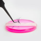 Close-up of tweezers holding eyelash extensions next to Platinum Adhesive on a pink platform for application.