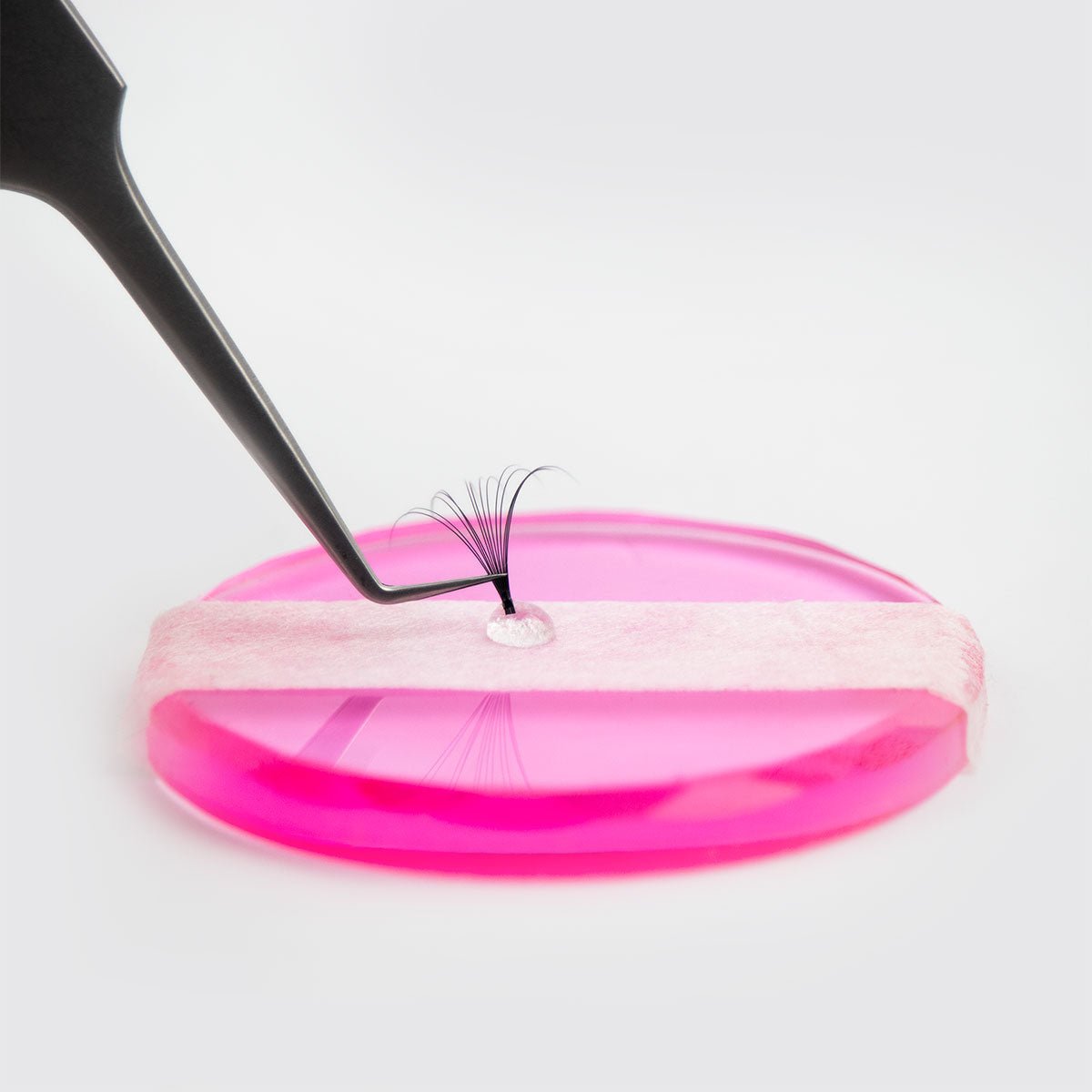 Close-up of tweezers holding eyelash extensions next to Platinum Adhesive on a pink platform for application.